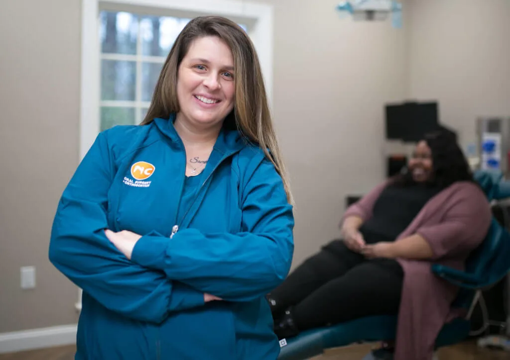 Oral Surgery Assistant smiling at camera with patient in turquoise dental chair behind her
