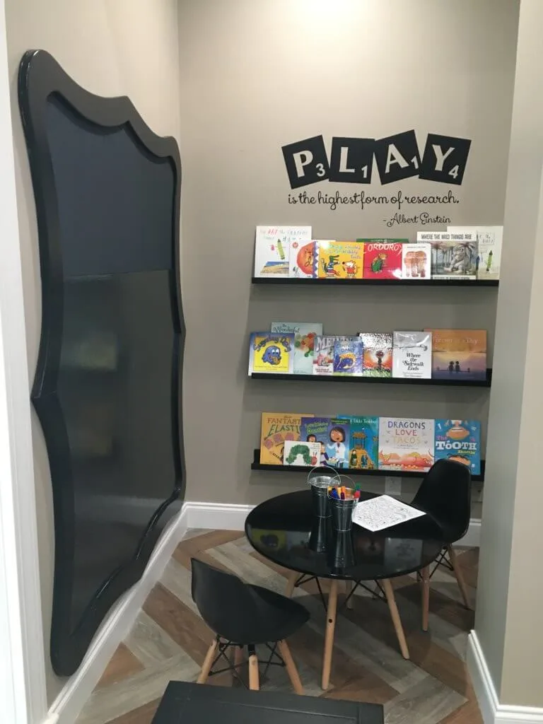 Kids corner with kids table, black dry erase board and markers, shelves filled with kids books, and PLAY written above shelf.